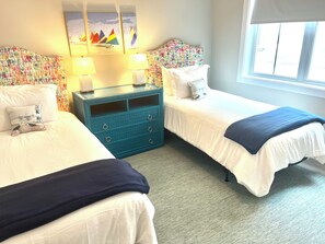 A hotel room with two beds, a blue dresser, and a window with blinds. - Shadow Brooke Village Unit 100 • FLETC & Vacation 3BR Condo w/Garage (St. Simons Island)
