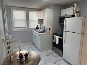 A compact kitchen with a dining area, featuring a refrigerator, stove, microwave, and a small sink. - 2-Bedroom Close to UNH & Portsmouth ~ Parking! (Dover)