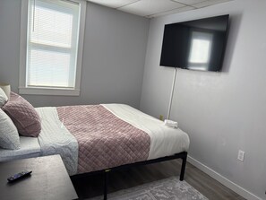 Ein Schlafzimmer mit einem Bett, einem an der Wand befestigten Fernseher, einem Fenster mit Jalousien und einer Fernbedienung auf einem kleinen Tisch.