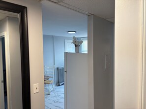 A hallway with a white door, a radiator, a white chair, and a table with a white vase on top. - 2-Bedroom Close to UNH & Portsmouth ~ Parking! (Dover)