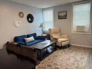 A living room with a blue sofa, a beige armchair, a coffee table, and a patterned rug. - 2-Bedroom Close to UNH & Portsmouth ~ Parking! (Dover)