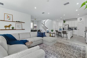 A modern living room with a sectional sofa, a dining area with a chandelier, and a kitchen with white cabinets. - 7BR Solara | Themed rooms, cinema, pool & spa (Kissimmee)