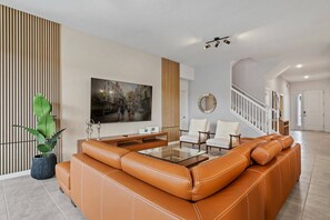 A spacious living room with a large brown leather sofa, a glass coffee table, and a painting on the wall. - 9BR themed home | Pool, arcade & cinema (Orlando)