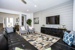 Un salon moderne avec un canapé en cuir noir, des fauteuils gris, un tapis à motifs et un téléviseur à écran plat fixé au mur.
