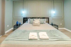 Una cama bien tendida con almohadas y toallas dobladas, una mesita de noche con una lámpara, y un aplique en la pared.