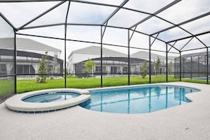 An indoor swimming pool with a hot tub, surrounded by a glass enclosure with a view of houses in the background. - 8BR themed home near Disney | Pool & game room (Kissimmee)