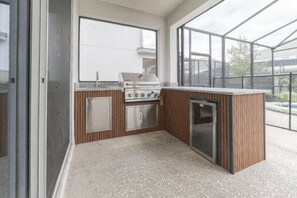 Cocina exterior compacta con estufa a gas, lavamanos y refrigerador, protegida por un muro de cristal.