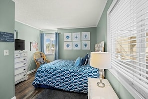 A bedroom with a bed, a dresser, a chair, a lamp, and a window with blinds. - Pelicans Watch 204- 3BR Steps From The Beach (Myrtle Beach)