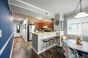 A compact kitchen with a central island, wooden bar stools, and a dining table with a patterned table runner. - Pelicans Watch 204- 3BR Steps From The Beach (Myrtle Beach)