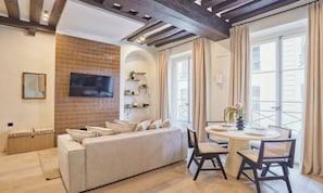 A living room with a sofa, a TV mounted on the wall, a dining table with chairs, and a balcony with curtains. - ELEGANT 2-BEDROOM MARAIS APARTMENT NEAR PLACE DES VOSGES (Paris)