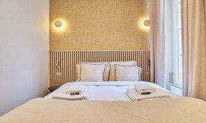 A hotel room with a bed, pillows, a headboard, and a window with curtains. - ELEGANT 2-BEDROOM MARAIS APARTMENT NEAR PLACE DES VOSGES (Paris)