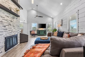 A spacious living area with a fireplace, a sofa, a television, and a dining area with a green cabinet. - NEW! Fall Hill Country | Hot Tub, Pool, Sleeps 11 (Dripping Springs)