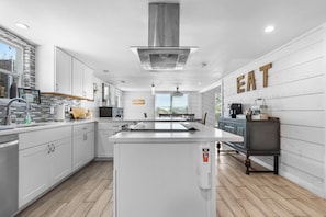 A modern kitchen with white cabinetry, a central island, and a 'EAT' sign on the wall. - NEW! Fall Hill Country | Hot Tub, Pool, Sleeps 11 (Dripping Springs)