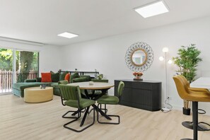 Ein modernes Wohnzimmer mit einem runden Tisch, grünen Stühlen, einem Sofa mit orangefarbenen und grünen Kissen, einer schwarzen Kommode und einem großen Spiegel an der Wand.