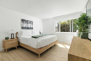 Ein modernes Schlafzimmer mit einem hölzernen Bett, weißer Bettwäsche, einem Nachttisch mit Lampe, einem Wandbild und einem großen Fenster mit Blick auf Grünflächen.