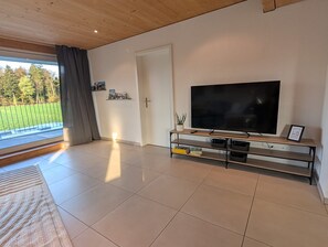 A spacious living room with a large flat-screen TV on a wooden TV stand, a door leading outside, and a view of a green landscape through a large window. - Waldblick Apartment, Quiet, Emmental, Solothurn & Bern (Kernenried)