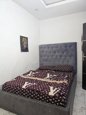 A bedroom with a grey upholstered headboard, a bed with a patterned bedspread, and a single wall-mounted artwork. - Spacious 2-Bedroom Escape | Stylish, Serene & Fully Equipped (Agege)
