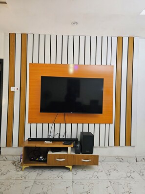 A flat-screen TV mounted on a wall with a wooden panel design, a wooden entertainment unit with a drawer below, and a marble-tiled floor. - Spacious 2-Bedroom Escape | Stylish, Serene & Fully Equipped (Agege)
