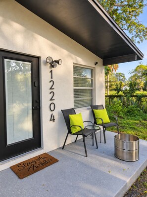 A modern house exterior with a black door, numbered 12204, a small window, and two chairs with green cushions on a concrete porch. - King Beds  Tampa Nature Hideaway Chic 2BR (Tampa)