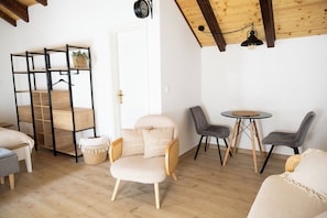 Ein Zimmer mit Holzboden, einer weißen Tür, einem schwarzen Metallregal, einem gepolsterten Sessel, zwei grauen Stühlen, einem kleinen runden Tisch und einer Pendelleuchte.