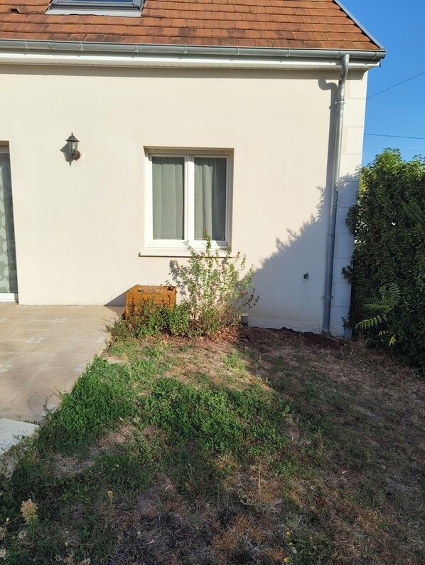 A house with a tiled roof, a window with white curtains, and a small garden area with a wooden planter and shrubs.