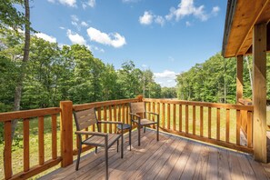 Eine Holzdielen-Terrasse mit zwei Stühlen und einem Tisch, umgeben von einem Geländer, mit Blick auf ein bewaldetes Gebiet.
