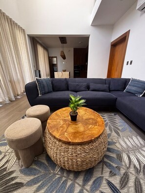 A modern living room with a dark sectional sofa, a round wooden coffee table with a plant, and two ottomans. - Brand-New 4-Bedroom Villa with Private Pool, SOV (Sosua)