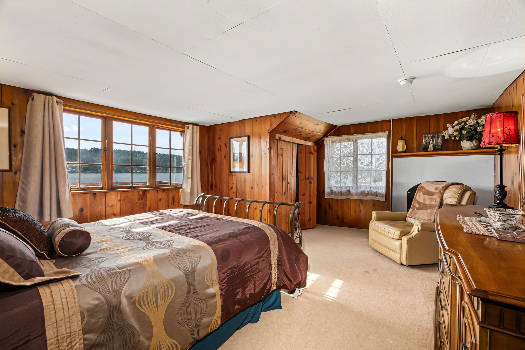 A bedroom with a large bed, a chair, a desk, and a view of a lake through the windows.