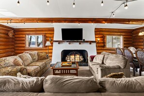 A log cabin interior with a fireplace, two sofas, a coffee table, and a television mounted above the fireplace.