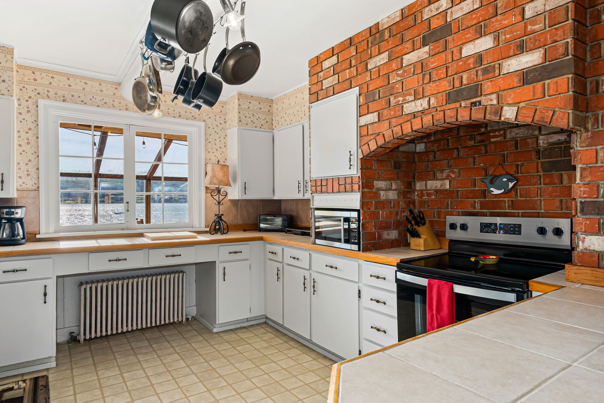 A kitchen with white cabinets, a brick wall, a window, and a hanging pot rack.