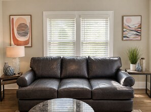 A dark brown leather sofa with a glass coffee table in front, positioned in a living room with white blinds, a lamp, and two framed artworks on the walls. - Solace on Sherwood | Old Town Duplex, Dogs Welcome (Fort Collins)