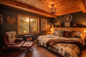 A cozy bedroom with a large bed, a leather armchair, a fireplace, and a view of the outdoors. - Luxury Cabin with EV Tesla Charger & Hot Tub! (Idyllwild-Pine Cove)