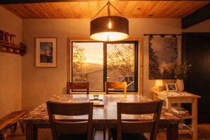 A dining area with a table and chairs, a pendant light, a window with a view, and a small table with a lamp and picture frame. - Luxury Cabin with EV Tesla Charger & Hot Tub! (Idyllwild-Pine Cove)