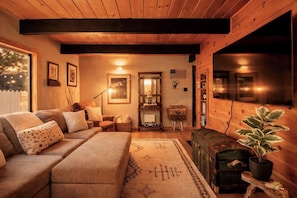 A cozy living room with a sofa, a lamp, a fireplace, and a television. - Luxury Cabin with EV Tesla Charger & Hot Tub! (Idyllwild-Pine Cove)