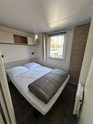 Una cama individual con cabecera, una ventana con cortinas, un armario de madera y una pequeña estantería sobre la cama.