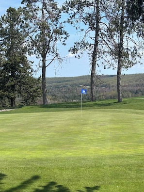A golf course with a flagstick on the green, surrounded by trees and a hilly landscape in the background.