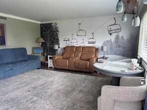 A living room with a brown sofa, a blue couch, and a grey armchair. There is a wall mural of ski lifts and a window with blinds.