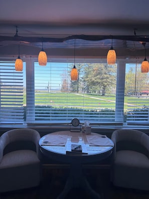 A room with a round table, two chairs, and a view of a park through large windows with blinds.