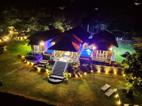 Una casa de dos plantas con piscina, mobiliario exterior y un jardín bien iluminado de noche.