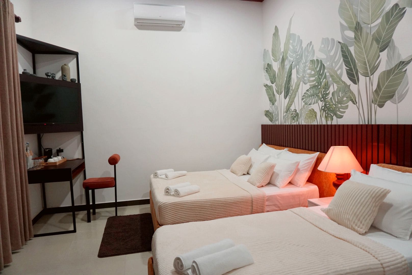 Una habitación de hotel con dos camas, un escritorio, una silla, un televisor y una pared decorada con un mural de plantas.