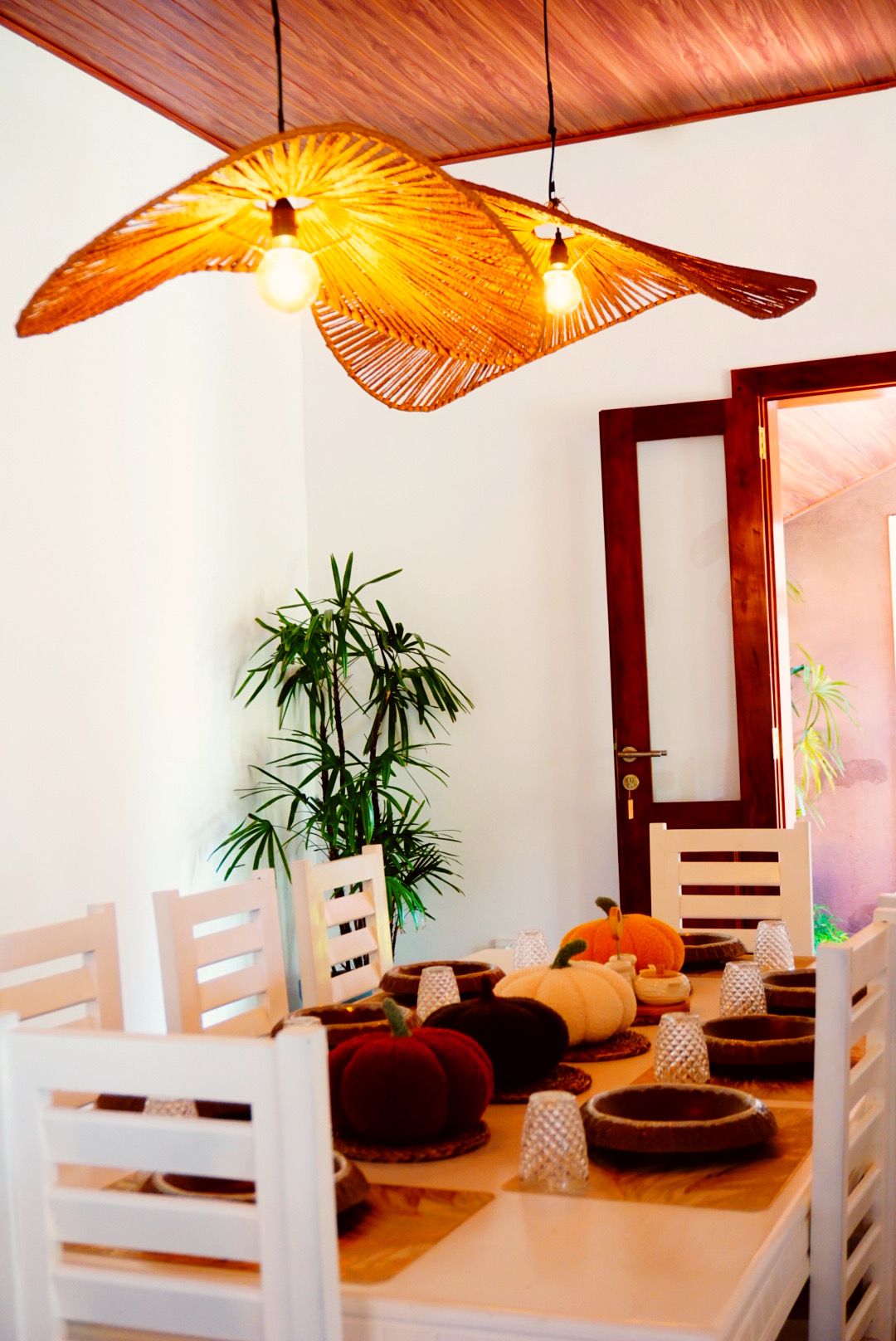 A dining area with a wooden ceiling, a white table, and chairs. There are decorative lights and a plant.