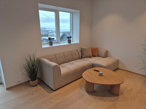Ein modernes Wohnzimmer mit einem beigefarbenen Ecksofa, einem runden Couchtisch und einem Fenster mit Blick auf Gebäude und ein Gewässer.