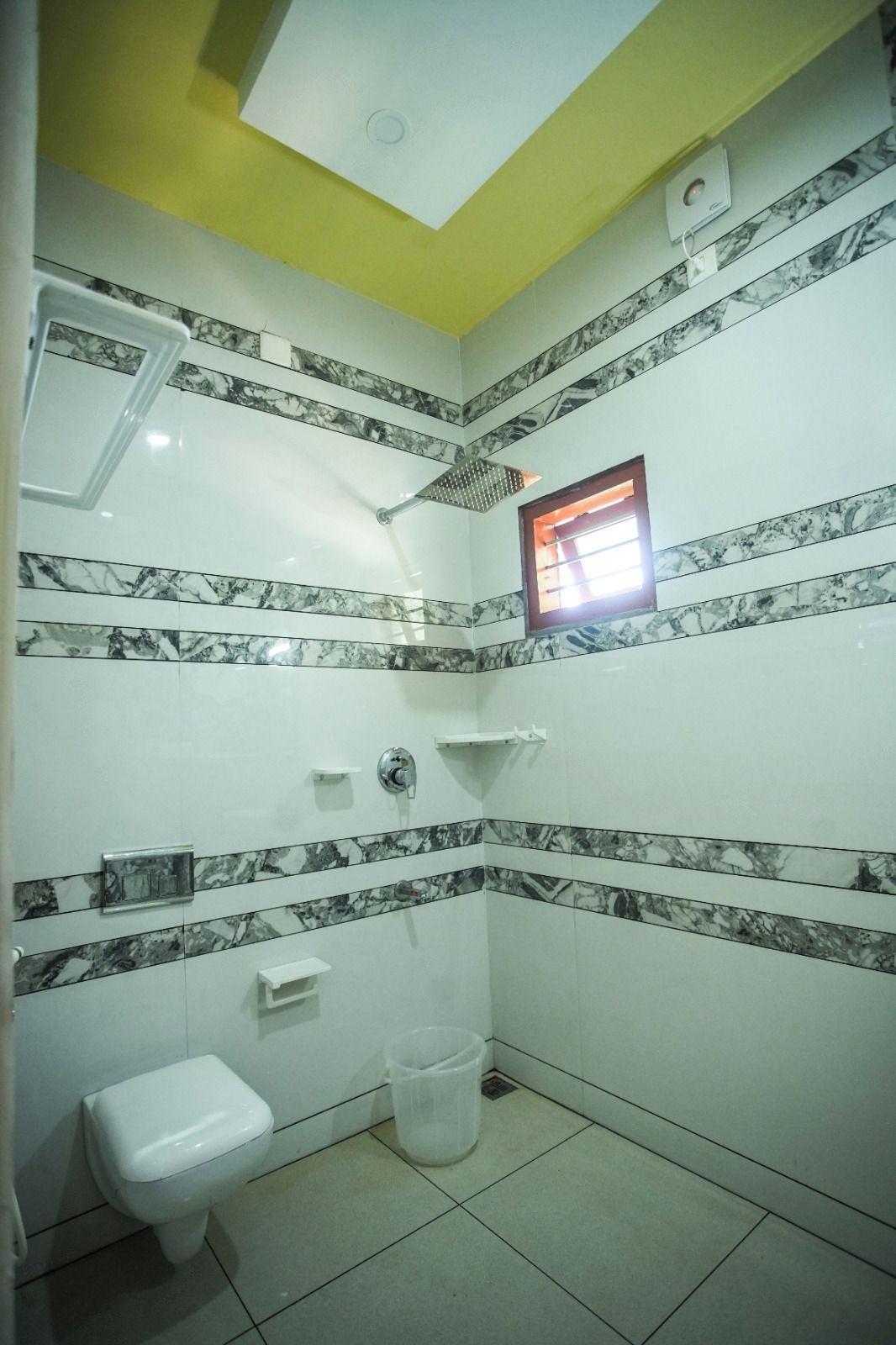 Un baño con baldosas de mármol blanco y negro, un inodoro blanco, una ducha con cabezal de ducha manual y una ventana pequeña con marco rojo.
