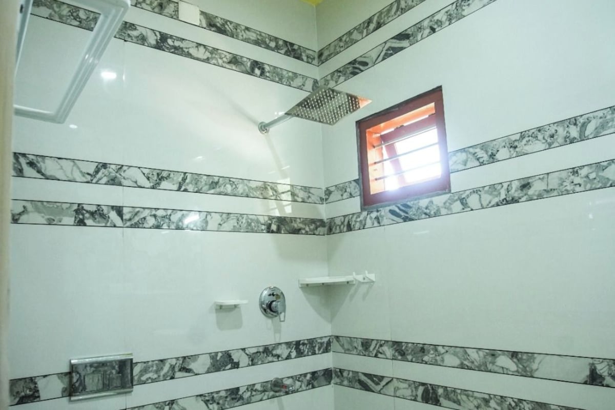 Un baño con baldosas de mármol blanco y negro, un inodoro blanco, una ducha con cabezal de ducha manual y una ventana pequeña con marco rojo.