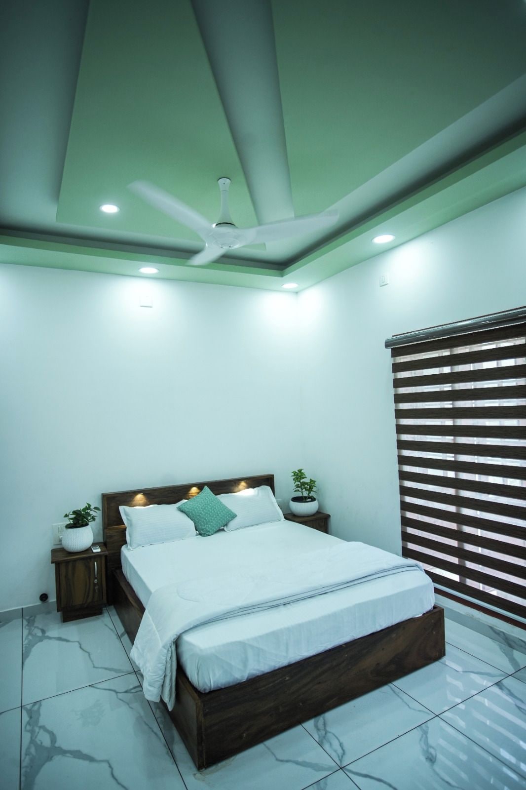 Une chambre à coucher avec un lit, des tables de chevet, un ventilateur de plafond et une fenêtre avec des stores.
