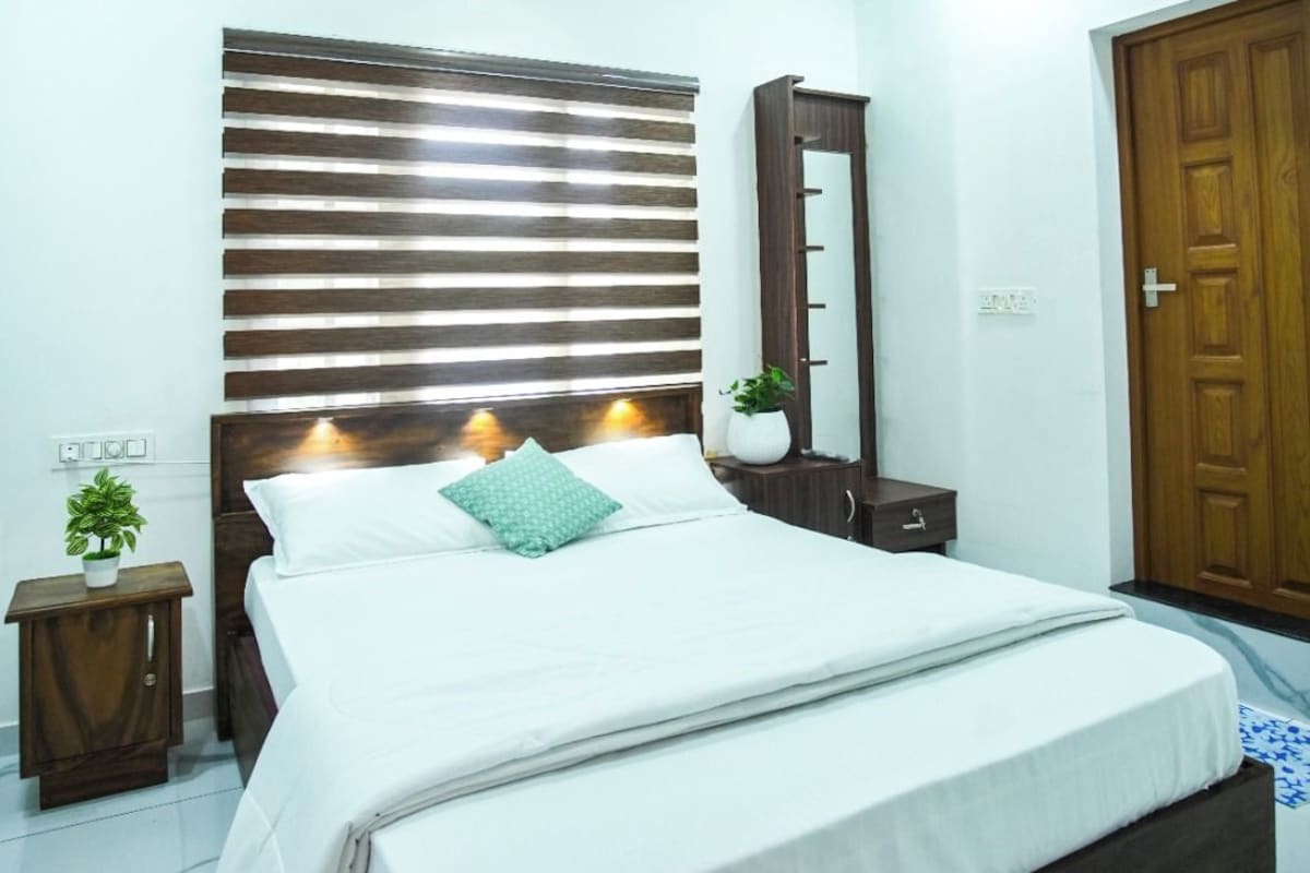 A bedroom with a bed, a ceiling fan, a wooden headboard, a bedside table with a plant, and a door.