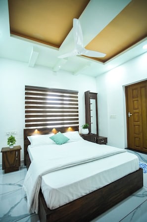Un dormitorio con cama, ventilador de techo, cabecera de madera, mesita de noche con planta y una puerta.
