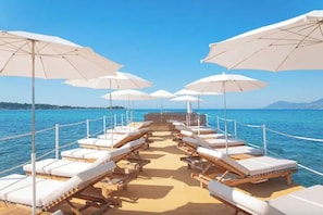 Een rij ligbedden met witte kussens en beige parasols op een terras met uitzicht op zee.