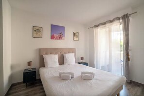 Ein ordentlich bezogenes Bett mit weißen Leinen und zwei Kissen, ein Nachttisch mit einer Lampe und ein Fenster mit durchsichtigen Vorhängen.