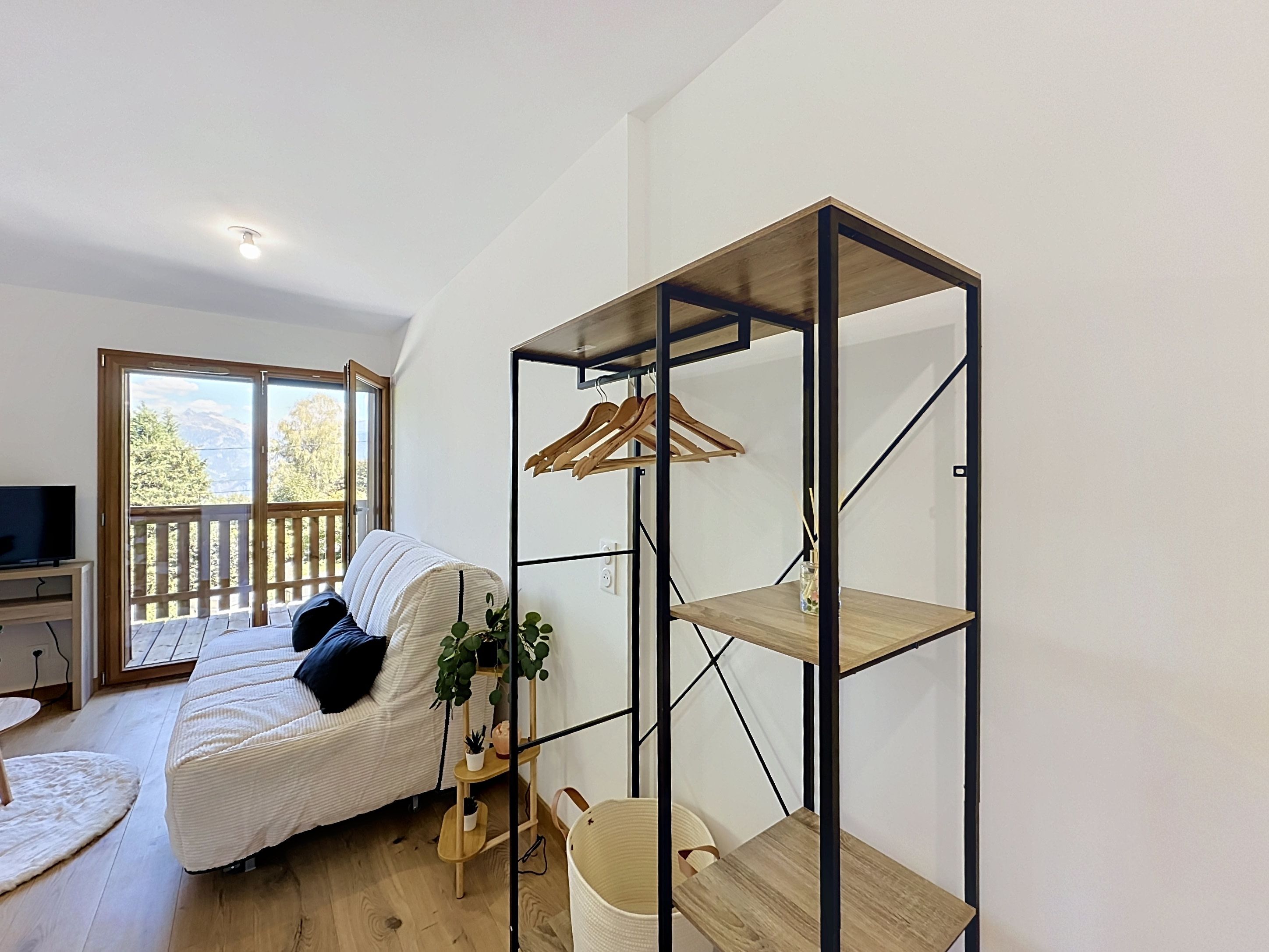 A modern living room with a sofa, a wooden floor, a TV stand, a balcony with a view of trees and mountains, and a metal shelving unit with a hanging rack.
